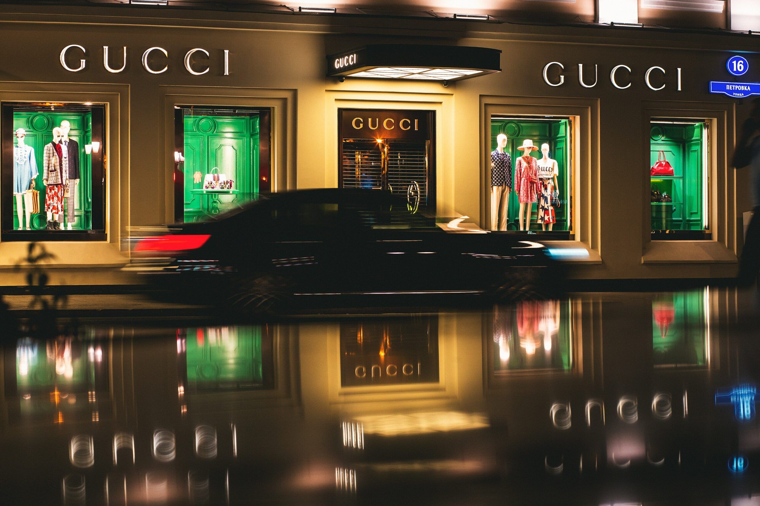 A Gucci store at night with green window displays and a black car driving past