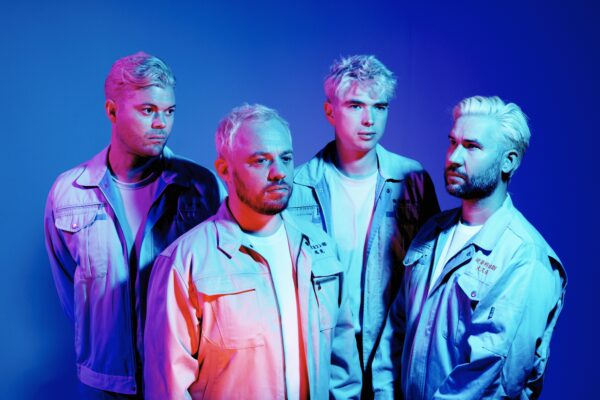 Four men in matching outfits stood against a purple a background. They are lit sharply with red and blue light.