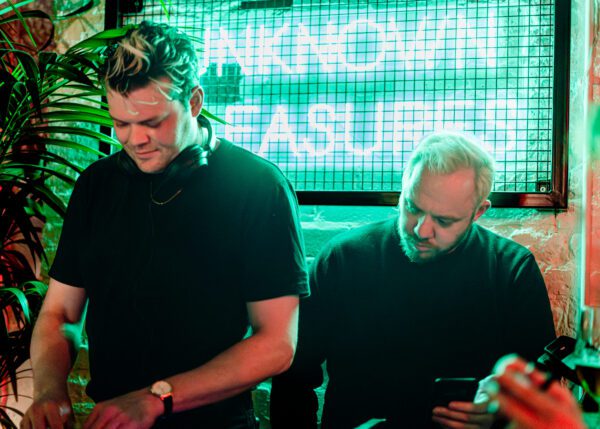 Jeremy Pritchard (left) and Jon Higgs (right) DJing in front of a green 'Unkown Pleasures' neon sign.