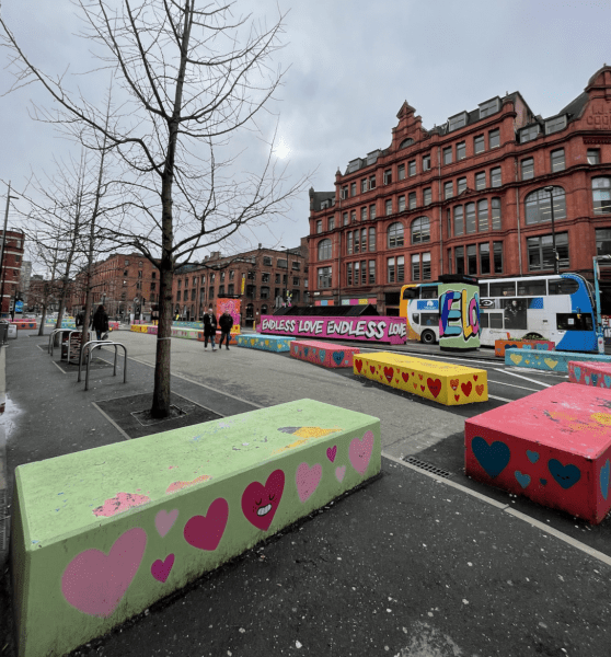 A photo of Stevenson Square with brightly coloured Hostile Vehicle Mitigation (HVM) Blocks that are decorated with graffiti hearts. The placement of the HVM blocks block a lot of the pavement. 