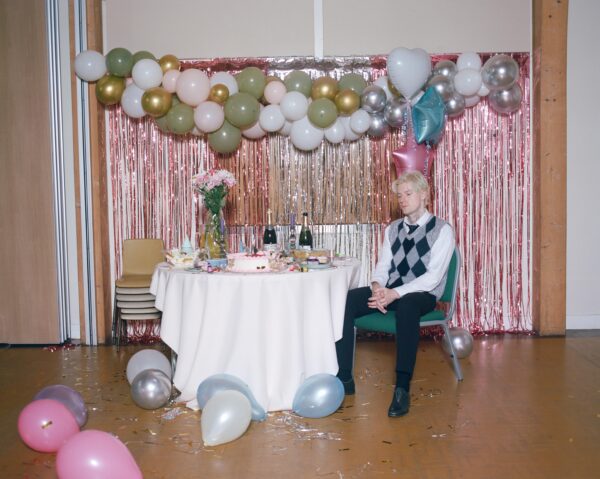 A man sat sadly at a small table in a village hall celebration scene. There are balloons above him and on the floor and a curtain of pink streamers behind him.