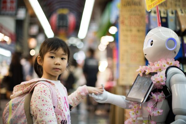 A young child holds the hand of a white robot wearing flowers around its neck
