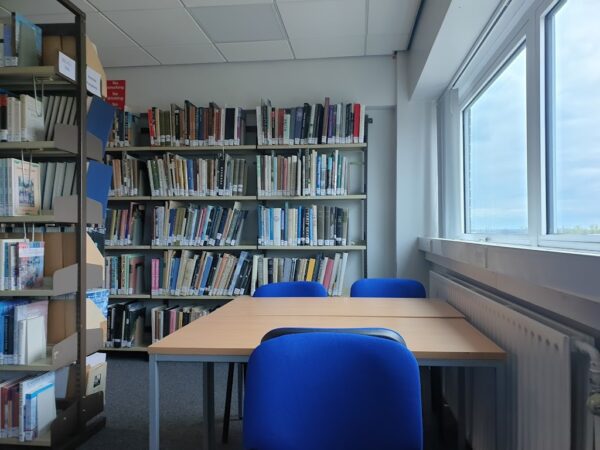 A photo depicting a library with some bookshelves, a table and three blue chairs.