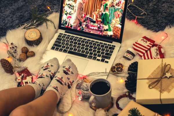 A laptop with a scene from Last Christmas on, someone wearing fuzzy socks surrounded by a hot drink and a present